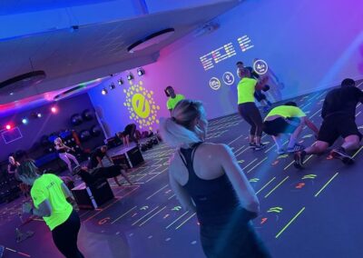 Bild på en dynamisk träningsklass hos Endorfin i form av Prama, med verksamhet i både Alingsås och Lerum. I ett modernt och välutrustat träningsrum med lila belysning och neonfärgade markeringar på golvet, utför deltagare intensiva stationsövningar såsom squats och planks. Endorfins karakteristiska logotyp lyser klart i bakgrunden, vilket förstärker varumärkets närvaro. Deltagarna är klädda i sportkläder, vissa med lysande neonfärger, vilket skapar en energisk och inspirerande träningsmiljö.